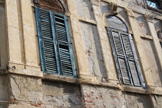 Weathered wooden shutters with peeling paint cover old, stately stone windows on a historical building facade. The architecture features detailed stonework with signs of age and wear, including cracks and exposed brick beneath the plaster.