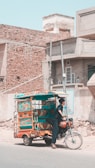 A vibrantly decorated auto rickshaw is parked on a street in front of rustic buildings with brick and concrete textures. A person dressed in dark traditional attire stands near the rickshaw, which has colorful designs featuring hearts and geometric patterns. The street is empty with some scattered construction material.
