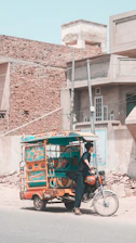A vibrantly decorated auto rickshaw is parked on a street in front of rustic buildings with brick and concrete textures. A person dressed in dark traditional attire stands near the rickshaw, which has colorful designs featuring hearts and geometric patterns. The street is empty with some scattered construction material.