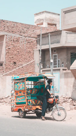 A vibrantly decorated auto rickshaw is parked on a street in front of rustic buildings with brick and concrete textures. A person dressed in dark traditional attire stands near the rickshaw, which has colorful designs featuring hearts and geometric patterns. The street is empty with some scattered construction material.
