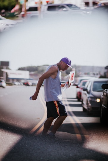 A person wearing a purple cap and casual clothing is standing on a road holding a sign with the letter 'H'. The scene features several parked cars along the road, and the atmosphere appears to be urban with an unfocused background.