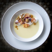 A gourmet dessert beautifully arranged in a white bowl, featuring creamy elements, caramelized popcorn, thinly sliced fruit, tiny cubes, and purple microgreens on a yellow sauce or custard. The plate is placed on another larger white plate, set against a textured surface.