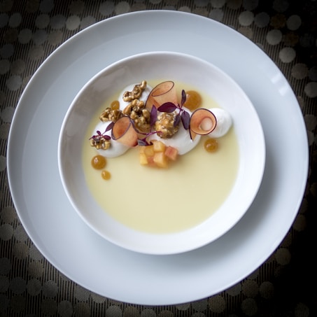A gourmet dessert beautifully arranged in a white bowl, featuring creamy elements, caramelized popcorn, thinly sliced fruit, tiny cubes, and purple microgreens on a yellow sauce or custard. The plate is placed on another larger white plate, set against a textured surface.