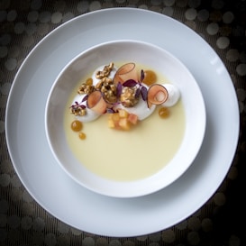 A gourmet dessert beautifully arranged in a white bowl, featuring creamy elements, caramelized popcorn, thinly sliced fruit, tiny cubes, and purple microgreens on a yellow sauce or custard. The plate is placed on another larger white plate, set against a textured surface.