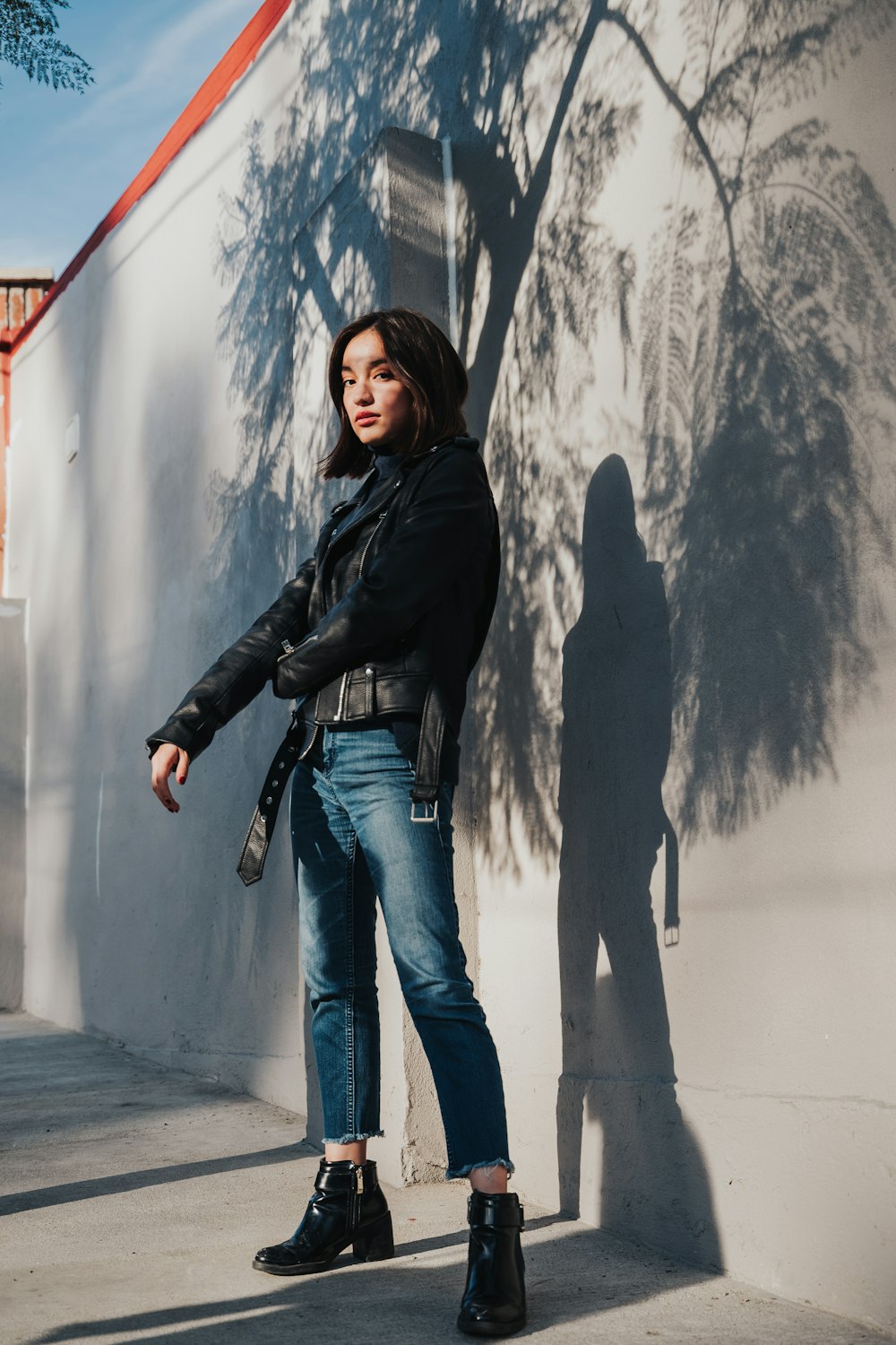 woman in black leather jacket and blue denim jeans standing beside white wall