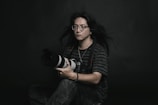 A person with long hair and glasses is holding a large camera. They are dressed in a striped shirt and sit against a dark, plain background. The mood is contemplative and artistic.
