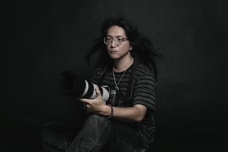 A person with long hair and glasses is holding a large camera. They are dressed in a striped shirt and sit against a dark, plain background. The mood is contemplative and artistic.