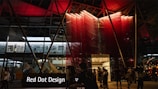 A modern building entrance illuminated at night with a striking red and black color scheme. People are mingling around, suggesting a social or event setting. The signage prominently displays 'Red Dot Design' in white letters against a black background.