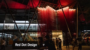 A modern building entrance illuminated at night with a striking red and black color scheme. People are mingling around, suggesting a social or event setting. The signage prominently displays 'Red Dot Design' in white letters against a black background.