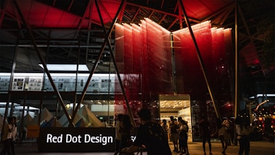 A modern building entrance illuminated at night with a striking red and black color scheme. People are mingling around, suggesting a social or event setting. The signage prominently displays 'Red Dot Design' in white letters against a black background.