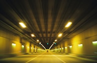 A long, illuminated tunnel with yellow and green lights creates a sense of speed and depth as the viewpoint appears to be moving through it. The ceiling and walls are lined with evenly spaced lights, and the road is empty except for a distant vehicle.