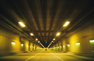 A long, illuminated tunnel with yellow and green lights creates a sense of speed and depth as the viewpoint appears to be moving through it. The ceiling and walls are lined with evenly spaced lights, and the road is empty except for a distant vehicle.