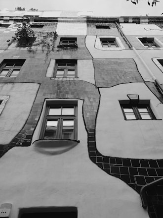 A building facade with an artistic, whimsical design featuring irregular windows and a wavy structure. The walls have a patchwork of textures and shades, and climbing plants add a touch of nature to the architecture. The lines and patterns create a sense of flow and creativity.