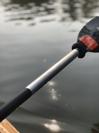 A close-up view of a kayak paddle partially submerged in a calm, reflective body of water. The paddle is held at an angle, with light reflections visible on the water's surface.