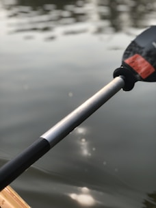 A close-up view of a kayak paddle partially submerged in a calm, reflective body of water. The paddle is held at an angle, with light reflections visible on the water's surface.