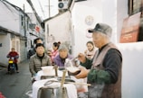 A street scene featuring several people gathered around a man serving hot food from large pots. The setting appears to be a narrow alley with traditional style buildings. Some people are eating while others look on. A motorcycle is visible in the background approaching the scene.