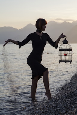 fine art photography,how to photograph a conceptual portrait; woman in black long sleeve dress holding white and black bird cage on water during daytime