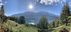 A serene landscape features a sunlit lake surrounded by lush greenery and snow-capped mountains. The foreground includes a sloping grass-covered area with trees and a small wooden structure. The bright sun creates a natural lens flare effect, and the sky is mostly clear with a few scattered clouds.