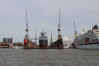 A large ship is docked at a shipyard, flanked by several cranes on either side. To the right, there's a large white cruise ship. The scene includes the backdrop of a cityscape with tall buildings. The water in the foreground reflects the industrial setting.
