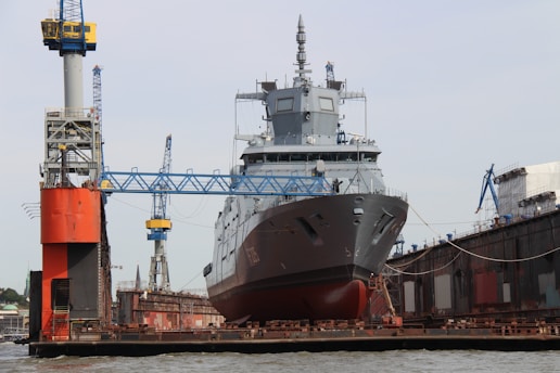 A large military ship is stationed in a dry dock, surrounded by cranes and industrial structures, indicating a shipbuilding or repair facility. The ship's hull is prominently visible with intricate detailing on its surface.