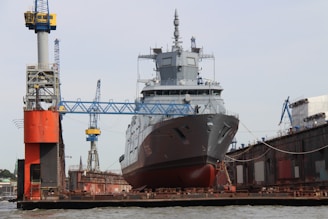A large military ship is stationed in a dry dock, surrounded by cranes and industrial structures, indicating a shipbuilding or repair facility. The ship's hull is prominently visible with intricate detailing on its surface.
