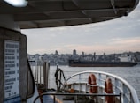 A large cargo ship labeled 'Dolce Vita' sails through a body of water. In the foreground, parts of another vessel are visible, including life rings and various ship components. A city skyline featuring numerous buildings and a prominent tower appears in the background under a cloudy sky.