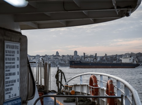 A large cargo ship labeled 'Dolce Vita' sails through a body of water. In the foreground, parts of another vessel are visible, including life rings and various ship components. A city skyline featuring numerous buildings and a prominent tower appears in the background under a cloudy sky.