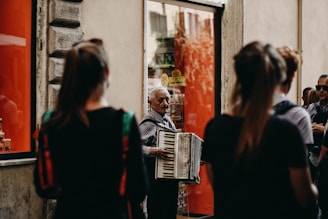 An older man is playing an accordion on a street, surrounded by several people. The background includes a large window with red panels and a stone wall. The people are all standing, and some seem to be watching the accordion player. The street has a somewhat urban, European feel to it.