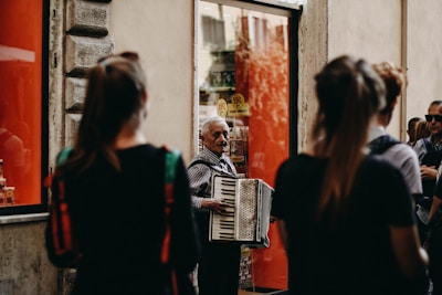 An older man is playing an accordion on a street, surrounded by several people. The background includes a large window with red panels and a stone wall. The people are all standing, and some seem to be watching the accordion player. The street has a somewhat urban, European feel to it.