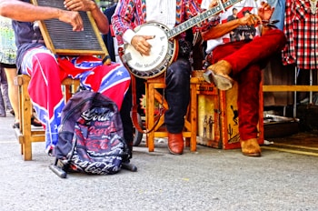 Three musicians sit on chairs, each holding an instrument. One plays a washboard, another a banjo, and the third appears to be clapping or using spoons. They wear colorful and casual attire, and a backpack with a distinct logo is placed on the ground.