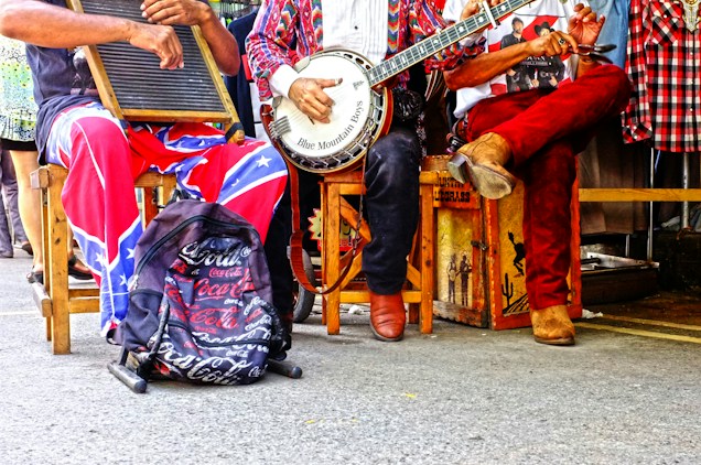 Three musicians sit on chairs, each holding an instrument. One plays a washboard, another a banjo, and the third appears to be clapping or using spoons. They wear colorful and casual attire, and a backpack with a distinct logo is placed on the ground.