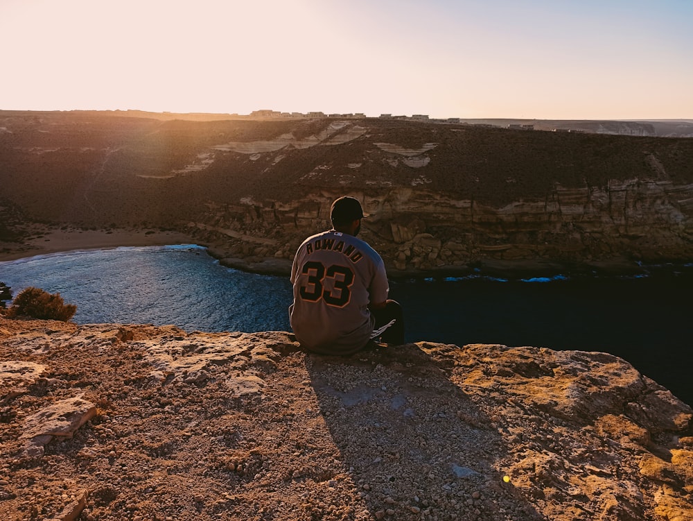 man in white hoodie sitting on brown rock near blue sea during daytime