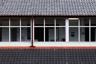 A building facade featuring a tiled roof and large rectangular windows. The windows reveal a glimpse of the interior, including walls and possible signage. The tiles on the roof are arranged in a neat, organized pattern, and there is a downspout running vertically between the windows.