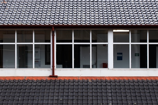 A building facade featuring a tiled roof and large rectangular windows. The windows reveal a glimpse of the interior, including walls and possible signage. The tiles on the roof are arranged in a neat, organized pattern, and there is a downspout running vertically between the windows.