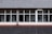 A building facade featuring a tiled roof and large rectangular windows. The windows reveal a glimpse of the interior, including walls and possible signage. The tiles on the roof are arranged in a neat, organized pattern, and there is a downspout running vertically between the windows.