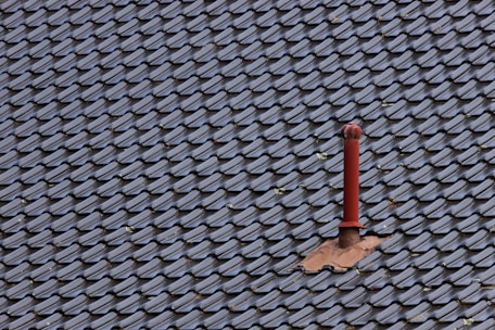 A roof covered with uniform, dark-colored tiles featuring a single red metal vent pipe protruding from the surface. Some tiles around the pipe are missing or displaced, revealing the underlying structure.