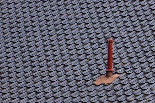 A roof covered with uniform, dark-colored tiles featuring a single red metal vent pipe protruding from the surface. Some tiles around the pipe are missing or displaced, revealing the underlying structure.