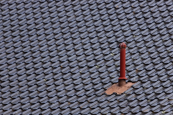 A roof covered with uniform, dark-colored tiles featuring a single red metal vent pipe protruding from the surface. Some tiles around the pipe are missing or displaced, revealing the underlying structure.