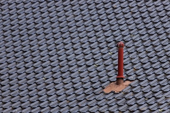 A roof covered with uniform, dark-colored tiles featuring a single red metal vent pipe protruding from the surface. Some tiles around the pipe are missing or displaced, revealing the underlying structure.