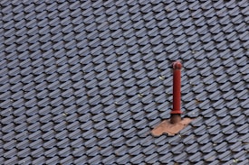 A roof covered with uniform, dark-colored tiles featuring a single red metal vent pipe protruding from the surface. Some tiles around the pipe are missing or displaced, revealing the underlying structure.