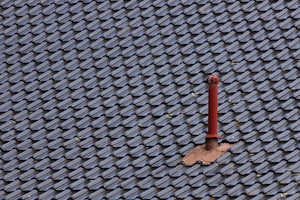 A roof covered with uniform, dark-colored tiles featuring a single red metal vent pipe protruding from the surface. Some tiles around the pipe are missing or displaced, revealing the underlying structure.