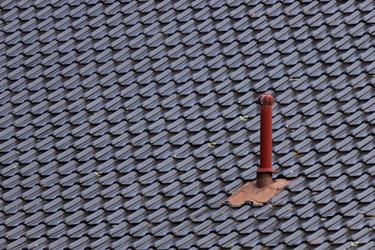 A roof covered with uniform, dark-colored tiles featuring a single red metal vent pipe protruding from the surface. Some tiles around the pipe are missing or displaced, revealing the underlying structure.