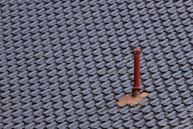A roof covered with uniform, dark-colored tiles featuring a single red metal vent pipe protruding from the surface. Some tiles around the pipe are missing or displaced, revealing the underlying structure.