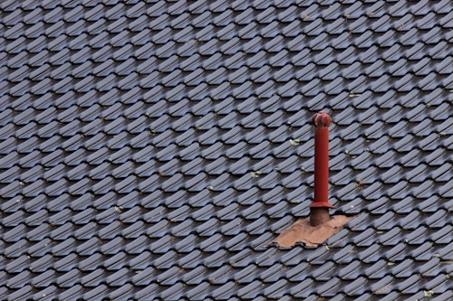 A roof covered with uniform, dark-colored tiles featuring a single red metal vent pipe protruding from the surface. Some tiles around the pipe are missing or displaced, revealing the underlying structure.