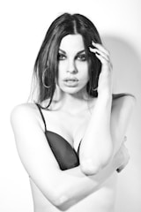 A black and white portrait of a person with long dark hair and prominent makeup. They are wearing a black bra and have an intense expression. The background is plain and the lighting creates strong contrasts.