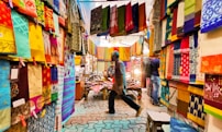 A vibrant textile market with colorful fabrics hanging from the walls and ceiling. The fabrics display a range of intricate patterns and designs. Two people walk through the market, one wearing a blue shirt and the other barely visible. The atmosphere seems lively and bustling, with soft lights illuminating the fabrics.