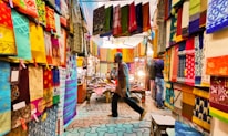 A vibrant textile market with colorful fabrics hanging from the walls and ceiling. The fabrics display a range of intricate patterns and designs. Two people walk through the market, one wearing a blue shirt and the other barely visible. The atmosphere seems lively and bustling, with soft lights illuminating the fabrics.