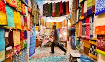 A vibrant textile market with colorful fabrics hanging from the walls and ceiling. The fabrics display a range of intricate patterns and designs. Two people walk through the market, one wearing a blue shirt and the other barely visible. The atmosphere seems lively and bustling, with soft lights illuminating the fabrics.