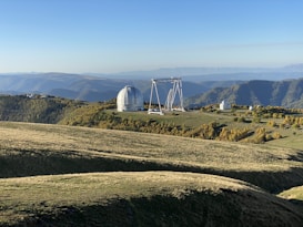 A large observatory with a dome and adjacent structures is situated on a hill surrounded by a lush green landscape and distant mountain ranges. The scene is bathed in sunlight, providing a clear view of the terrain and the observatory.