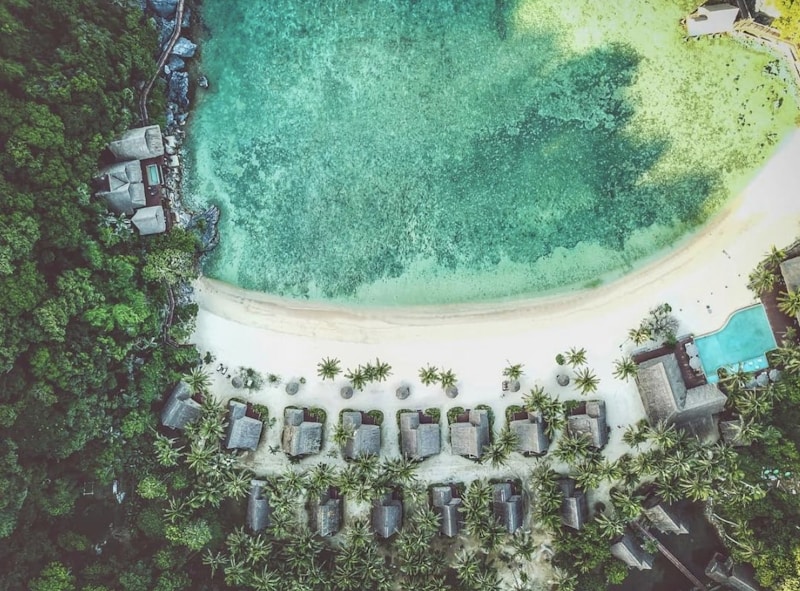 Aerial view of a tropical beach resort surrounded by lush greenery. Small villas are lined up along a pristine white sand beach, flanked by palm trees. The clear turquoise water of the sea gently laps the shoreline, providing a serene backdrop. A swimming pool is visible on the right, adding to the luxurious setting.